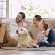 Rodzina siedząca razem w salonie – uśmiechnięty ojciec robi selfie z partnerką, małym dzieckiem i psem. Obrazek przedstawia ciepły, rodzinny moment, który wiele osób publikuje w mediach społecznościowych. Ilustracja może symbolizować sharenting – czyli dzielenie się wizerunkiem dziecka w internecie, często bez refleksji nad jego prywatnością i zgodą.