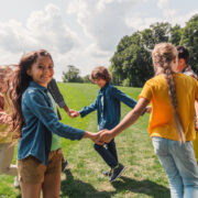 Grupa uśmiechniętych dzieci, trzymających się za ręce, bawi się radośnie na zielonej trawie w słoneczny dzień, celebrując beztroskę Dnia Dziecka
