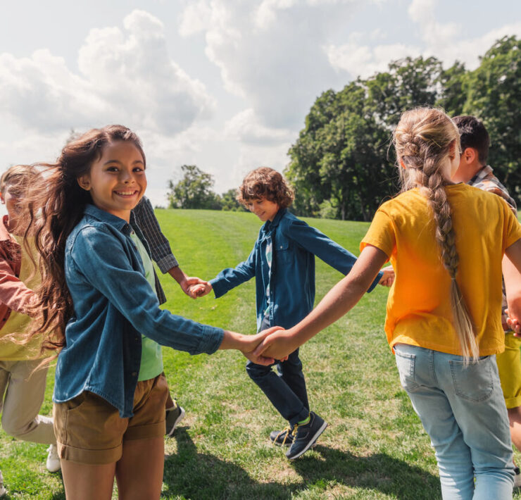 Grupa uśmiechniętych dzieci, trzymających się za ręce, bawi się radośnie na zielonej trawie w słoneczny dzień, celebrując beztroskę Dnia Dziecka