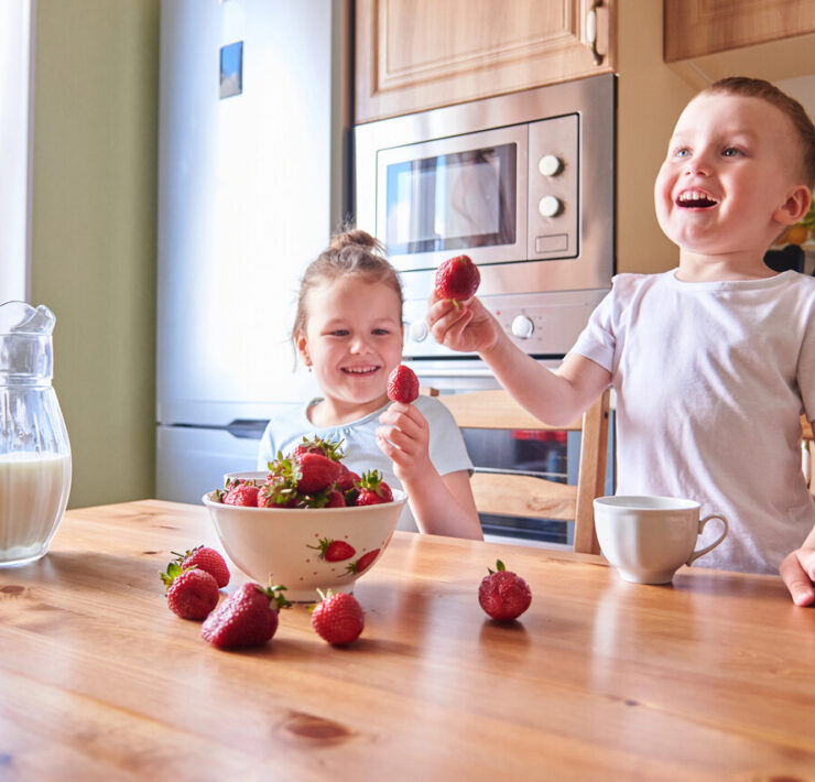 Dwoje małych dzieci siedzi przy drewnianym stole w jasnej kuchni. Chłopiec po prawej stronie trzyma w górze truskawkę i śmieje się, patrząc w lewo. Dziewczynka po lewej stronie trzyma truskawkę przy ustach, uśmiechając się i patrząc na chłopca. Na stole przed nimi stoi biała miska pełna truskawek, kilka pojedynczych truskawek, szklany dzbanek z mlekiem i biały kubek. W tle widać lodówkę, kuchenkę mikrofalową i fragment ściany z kafelkami.