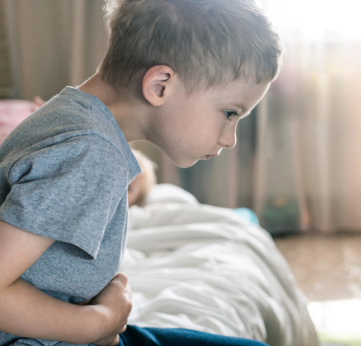 Chłopiec siedzi na łóżku, trzymając się za brzuch i spoglądając w dół z wyrazem dyskomfortu na twarzy.