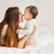 Uśmiechnięta młoda kobieta z długimi brązowymi włosami siedzi na białym łóżku i trzyma małe dziecko w białym ubranku. Ich twarze są blisko siebie, jakby miały się pocałować w nos. W tle widać miękkie, rozproszone światło przypominające lampki choinkowe.