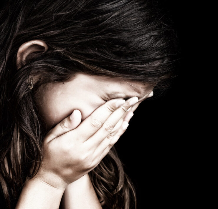 Zbliżenie przedstawia osobę z długimi, ciemnymi włosami, która zakrywa twarz obiema dłońmi. Głowa osoby jest lekko pochylona w dół i w prawo, a łokcie spoczywają na czymś, co wydaje się być nogami lub inną powierzchnią poza kadrem. Jej karnacja jest jasna, a w lewym płatku ucha widać mały złoty kolczyk. Tło jest jednolicie czarne, co w całości skupia uwagę na tej osobie. Oświetlenie jest przyciemnione i nieco dramatyczne, rzucając cienie na ramiona i dłonie, co podkreśla nastrój przygnębienia lub smutku. Ogólna kompozycja jest ciasna, koncentrując się na dłoniach zasłaniających twarz, co uniemożliwia zobaczenie bezpośredniej ekspresji emocjonalnej, pozostawiając widzowi interpretację postawy i gestu.