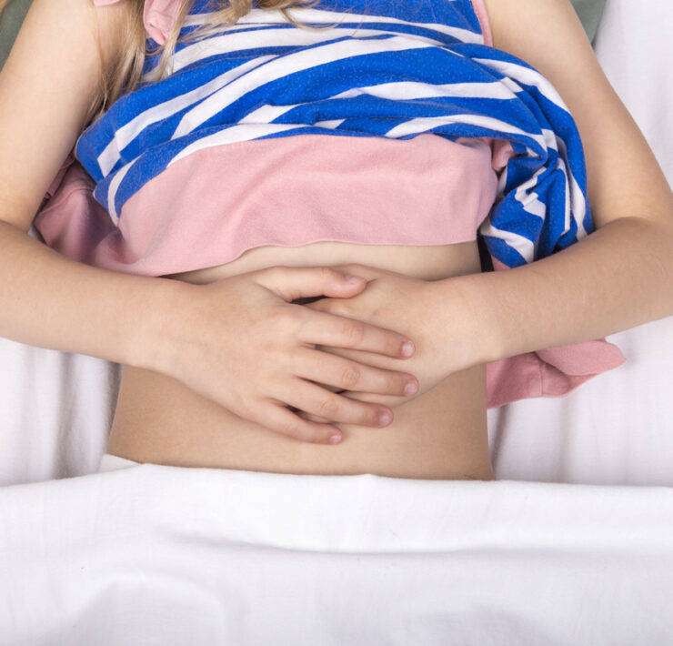 Ujęcie z góry przedstawia brzuch dziecka z objawem zapalenia wyrostka. Dziecko leży w łóżku pod białą kołdrą. Ma na sobie różową koszulkę i niebiesko-białą bluzkę w paski, częściowo podwiniętą, odsłaniającą brzuch. Dłonie są splecione i położone na brzuchu, co może sugerować ból lub dyskomfort w okolicy brzucha. W górnej części kadru widać fragmenty włosów i poduszki.