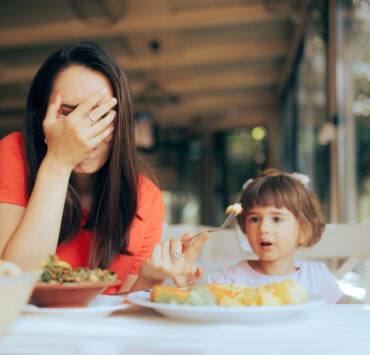Zmęczona kobieta siedzi przy stole z ręką zasłaniającą twarz, obok niej mała dziewczynka rozmawia i patrzy na nią, podczas gdy kobieta trzyma widelec z jedzeniem. Na stole stoją talerze z obiadem.