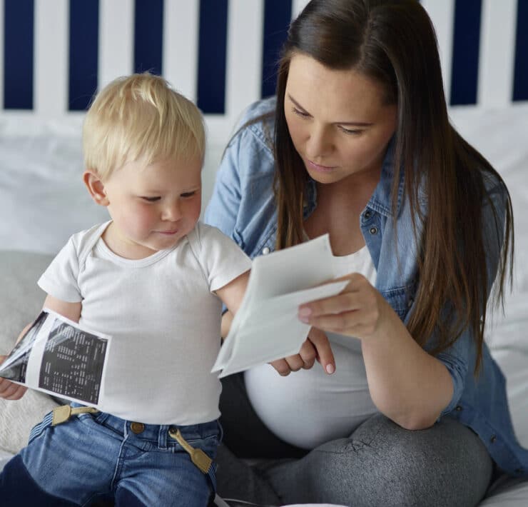 Na zdjęciu widać kobietę w ciąży z długimi, ciemnymi włosami, ubraną w jeansową koszulę i szare legginsy. Siedzi na łóżku obok małego dziecka z blond włosami, ubranego w białą koszulkę i jeansowe spodenki. Oboje skupiają się na oglądaniu zdjęć USG. Kobieta trzyma jedno zdjęcie, a dziecko drugie, uważnie się im przyglądając. W tle widać białe poduszki i wezgłowie łóżka w biało-granatowe pasy.