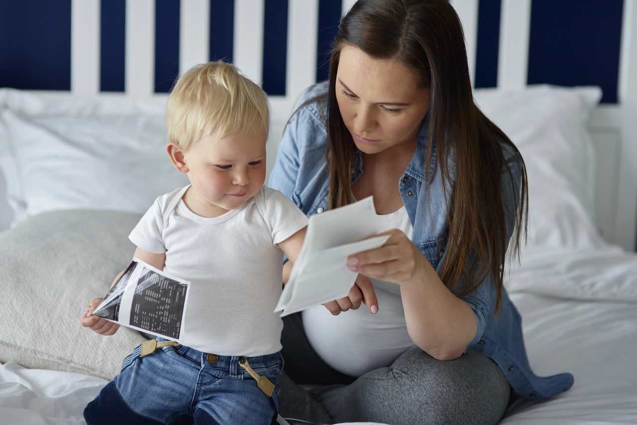 Na zdjęciu widać kobietę w ciąży z długimi, ciemnymi włosami, ubraną w jeansową koszulę i szare legginsy. Siedzi na łóżku obok małego dziecka z blond włosami, ubranego w białą koszulkę i jeansowe spodenki. Oboje skupiają się na oglądaniu zdjęć USG. Kobieta trzyma jedno zdjęcie, a dziecko drugie, uważnie się im przyglądając. W tle widać białe poduszki i wezgłowie łóżka w biało-granatowe pasy.