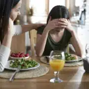 Nastolatka z zaburzeniami odżywiania z głową ukrytą w dłoniach siedzi przy stole, obok niej dorosła kobieta kładzie wspierająco rękę na jej ramieniu. Na stole leżą talerze z sałatką i szklanki z napojami. Atmosfera sugeruje trudną rozmowę związaną z jedzeniem