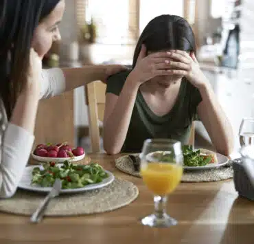 Nastolatka z zaburzeniami odżywiania z głową ukrytą w dłoniach siedzi przy stole, obok niej dorosła kobieta kładzie wspierająco rękę na jej ramieniu. Na stole leżą talerze z sałatką i szklanki z napojami. Atmosfera sugeruje trudną rozmowę związaną z jedzeniem
