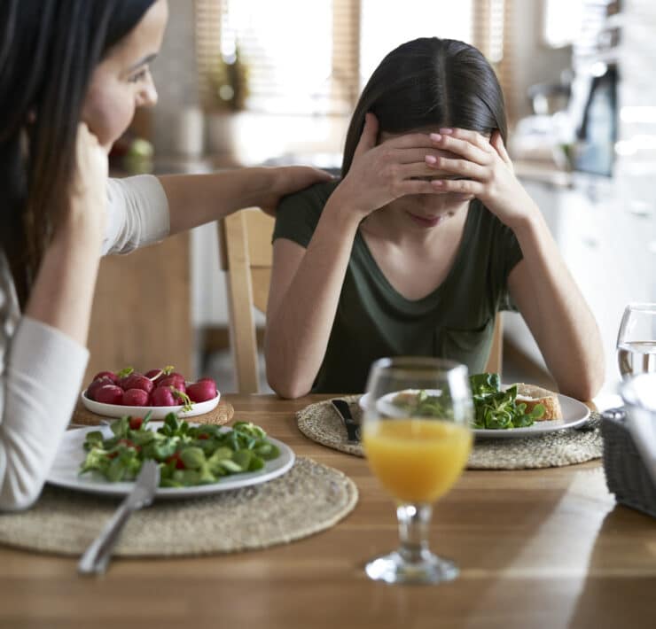 Nastolatka z zaburzeniami odżywiania z głową ukrytą w dłoniach siedzi przy stole, obok niej dorosła kobieta kładzie wspierająco rękę na jej ramieniu. Na stole leżą talerze z sałatką i szklanki z napojami. Atmosfera sugeruje trudną rozmowę związaną z jedzeniem