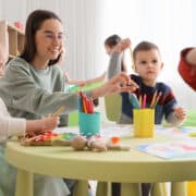 Dzieci w przedszkolu podczas zajęć plastycznych z nauczycielką. Wyprawka do przedszkola obejmuje przybory plastyczne, kredki i inne kreatywne akcesoria potrzebne na zajęciach.