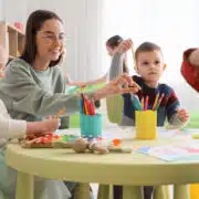 Dzieci w przedszkolu podczas zajęć plastycznych z nauczycielką. Wyprawka do przedszkola obejmuje przybory plastyczne, kredki i inne kreatywne akcesoria potrzebne na zajęciach.