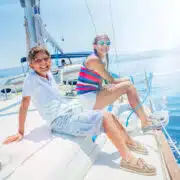 Młody chłopiec i dziewczynka siedzą na pokładzie żaglówki, uśmiechając się do aparatu. Chłopiec na pierwszym planie ma na sobie białą koszulkę polo i szorty w paski. Dziewczynka za nim ubrana jest w kolorowy, pasiasty top na ramiączkach i białe szorty. Oboje cieszą się słonecznym dniem na wodzie, a w prawym górnym rogu zdjęcia widać odblask słońca